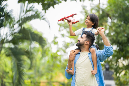 Happy smiling indian girl kid playing with airplane toy by sitting on father shoulder at park - concept of freedom, family support and togetherness