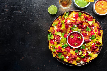 Loaded nachos. Mexican nacho chips with beef, overhead flat lay shot with guacamole sauce, cheese salsa, tequila drinks, limes, on a black slate background with copy space - obrazy, fototapety, plakaty