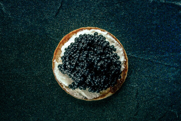 A blini with caviar and cream cheese, overhead flat lay shot on a black slate background