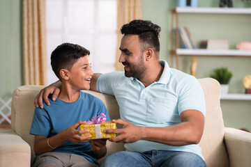 Excited indian kid receiving surprise gift from father while sitting on sofa at home - concept of...