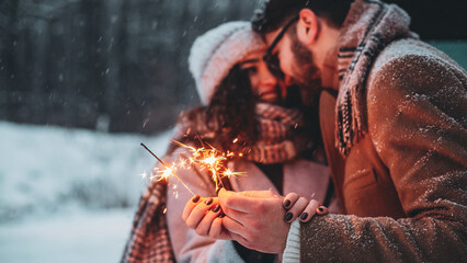 Outdoor waist up portrait of young beautiful happy smiling couple posing on street. Models hugging, looking at each other, holding sparkles, wearing stylish clothes. Snowfall. Copy, empty space - obrazy, fototapety, plakaty