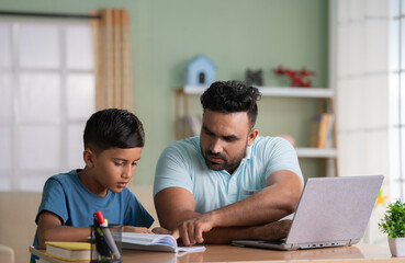 Indian father helping his son for reading while working on laptop at home - concept of Educational...