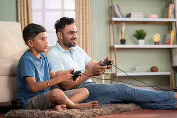 Happy excited indian father with son playing video game using joystick on floor at home - concept of Joyful playtime, weekdn fun and relationship bonding