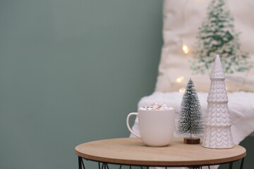 Christmas home decorations - cup of cocao with marshmellow, ceramic christmas tree on coffee table on green background.