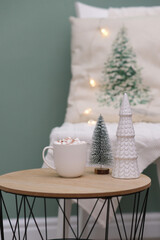 Christmas home decorations - cup of cocao with marshmellow, ceramic christmas tree on coffee table on green background.