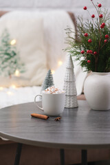 cup of cacao with marshmellows, christmas tree, branches fir in vase on coffee table on white pillow background.