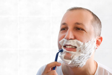A young man shaves with a disposable razor in the bathroom while looking in the mirror. Health,...