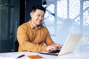 Successful asian programmer developer working with laptop inside office, man in shirt smiling and typing on keyboard, coding new software, businessman happy with achievement.