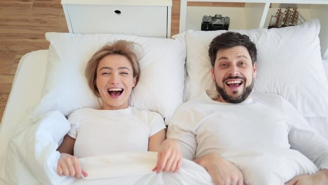 Top view of loving happy caucasian couple woke up while lying on white pillows under the covers on soft white bed in modern apartment.