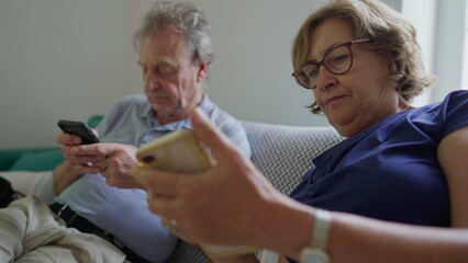 Obraz premium Senior couple looking in Their Phones seated on couch staring at screen, Older married man and woman using modern technology, holding smartphone