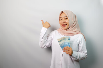Smiling young Asian hijab woman holding paper money and pointing at banner advertisement standing over white background.