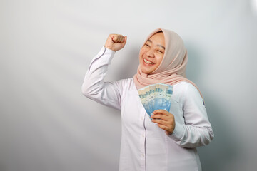 Excited Asian Muslim woman clenched fist while holding paper money and showing happy expression isolated on white background
