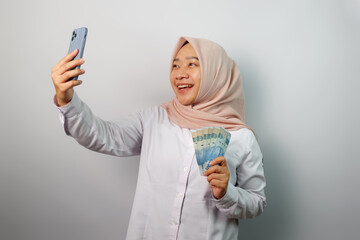 Shocked young Asian hijab woman using mobile phone and holding money banknotes isolated on white background