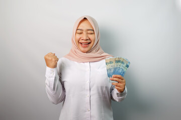 Excited Asian Muslim woman holding paper money (Indonesian rupiah banknotes) and celebrating luck isolated on white background.
