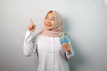 Smiling young Asian hijab woman holding paper money and pointing at banner advertisement standing over white background.