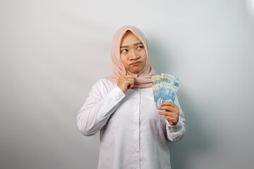 Portrait of cute Asian hijab standing isolated over white background holding cash in her hand thinking by looking at the copy space.