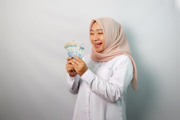 Enthusiastic Asian hijab woman looking excited at camera and holding money rupiah in hand isolated on white background