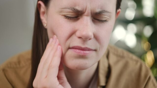 Close up of unhappy sad young woman hand touching cheek sitting on couch, face expression from toothache, sensitivity, having tooth or teeth problem, suffering from health. Sensitive teeth concept