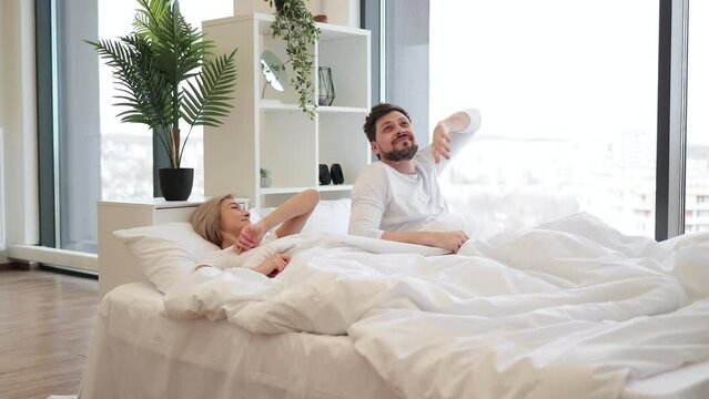 Loving caucasian couple in casual attire stretching their limbs after night sleep while still lying in bed of bright bedroom. Relaxed family man and woman starting new sunny day in modern flat.