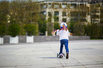 Adorable preschooler girl riding her scooter in a city park on sunny spring day