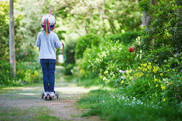 Adorable preschooler girl riding her scooter in a city park on sunny spring day - obrazy, fototapety, plakaty