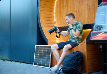 Excited photographer looks at the photos he just took. Young photographer sitting and checking result of photosession. Handsome male sitting on round bench with solar panel on background.