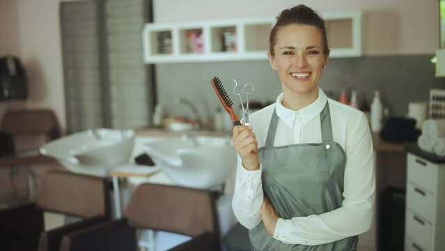 happy 40 years old woman hairdresser in modern beauty salon in apron with scissors near salon backwash unit.