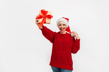 Photo of a smiling woman wearing a Santa hat and a red knitted sweater, holding a small gift box for New Year's celebration.