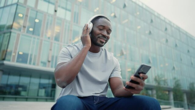 African american man music lover in headphones enjoying listening music on smartphone sitting on city street outdoors. Positive smiling guy melomaniac rhythmically nodding head relaxing moving body.