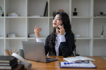 Freelance Asian woman talking on the phone with friend or colleague while working on laptop