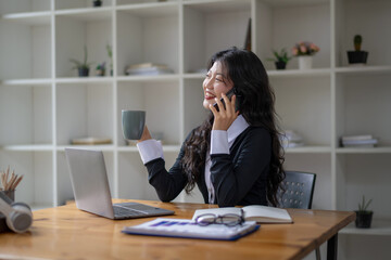 Freelance Asian woman talking on the phone and sipping coffee while working at the desk.