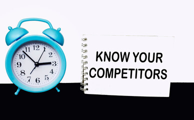 View of a notepad with the text KNOW YOUR COMPETITORS, clock on a black and white background. Business concept.
