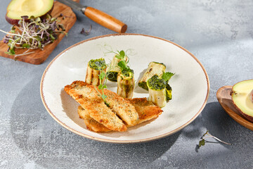 Horizontal side view of tempura perch fillet served with zucchini rolls on a ceramic plate. The grey concrete background and harsh shadows accentuate the fine details of this gourmet dish