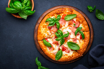 Homemade hot pizza with smoked ham, cheese, spicy tomato sauce and green basil on black table background, top view