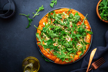 Hot homemade pizza with blue cheese, ham, pesto sauce and fresh arugula, black table background, top view