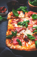 Rectangular hot pizza with ham, cheese, dried tomatoes, spicy sauce and basil, black table background, top view