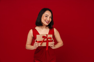 Fototapeta premium Happy woman holding gift box while standing isolated over red background
