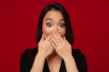 Beautiful asian woman covering her mouth while standing isolated over red background