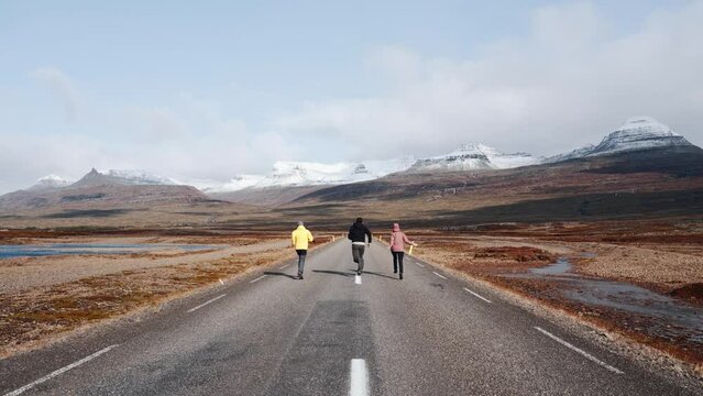 Three people run in the middle of the street. Friends run happily along a long lonely road with snow-capped mountains in the background. Idyllic image of freedom, youth and hiking. Travel and explore.