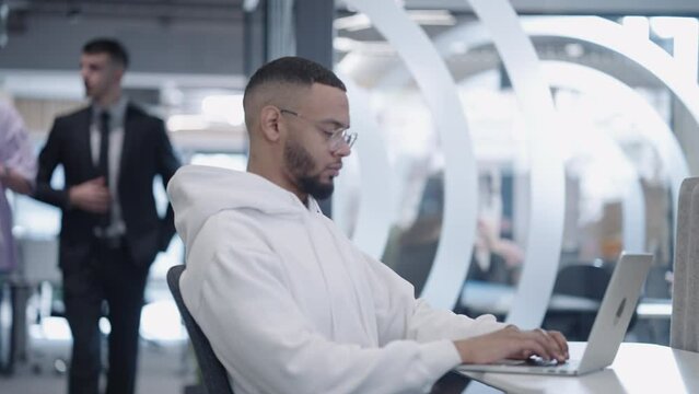 African-American man works diligently on his laptop in a modern office environment while his diverse colleagues move around him, depicting a focused and productive professional workspace
