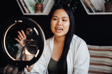 Asian woman making video content for social media with smartphone and ring lamp while sitting at home