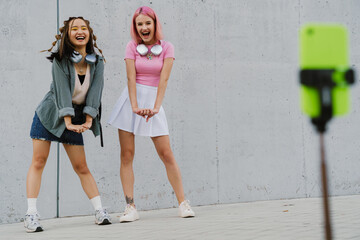 Two joyful girls making videos with smartphone and dancing while standing outdoors near gray wall