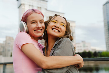 Two smiling girls hugging while standing at city street - obrazy, fototapety, plakaty