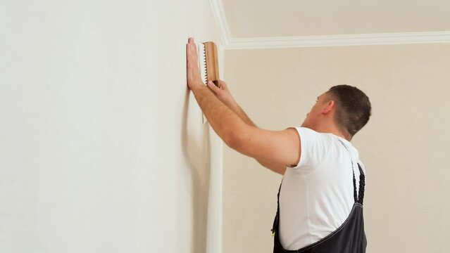 Vinyl wallpapers squeegee tool in use by professional interior renovation worker. Young man with dark hair in a uniform glueing beige wallpaper at home.