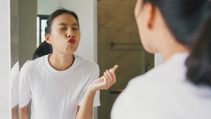 Young Asian happy woman make up looking at her reflection in mirror apply lip balm on her lips in...