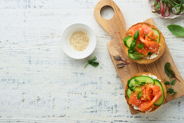 Open smoked salmon sandwiches with cream cheese, cucumber, sesame seeds, microgreens, spinach, and peas leaves on light old wooden background. Healthy breakfast food. Delicious snack. Top view.