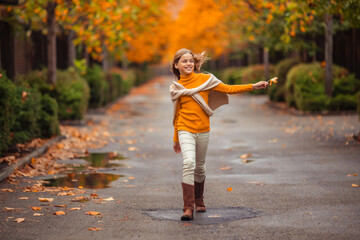 Fototapeta premium a teenage girl in a yellow sweater walks along a street outside the city against the backdrop of yellow trees. fun walk in autumn