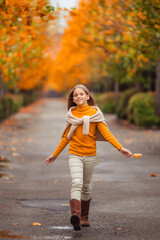 a teenage girl in a yellow sweater walks along a street outside the city against the backdrop of yellow trees. fun walk in autumn