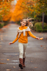 a teenage girl in a yellow sweater walks along a street outside the city against the backdrop of yellow trees. fun walk in autumn