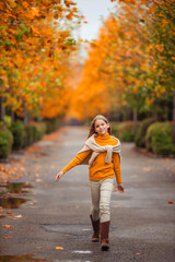a teenage girl in a yellow sweater walks along a street outside the city against the backdrop of yellow trees. fun walk in autumn